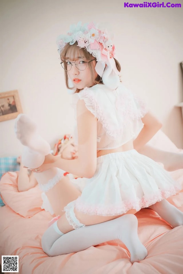 A woman in a white dress and glasses sitting on a bed.